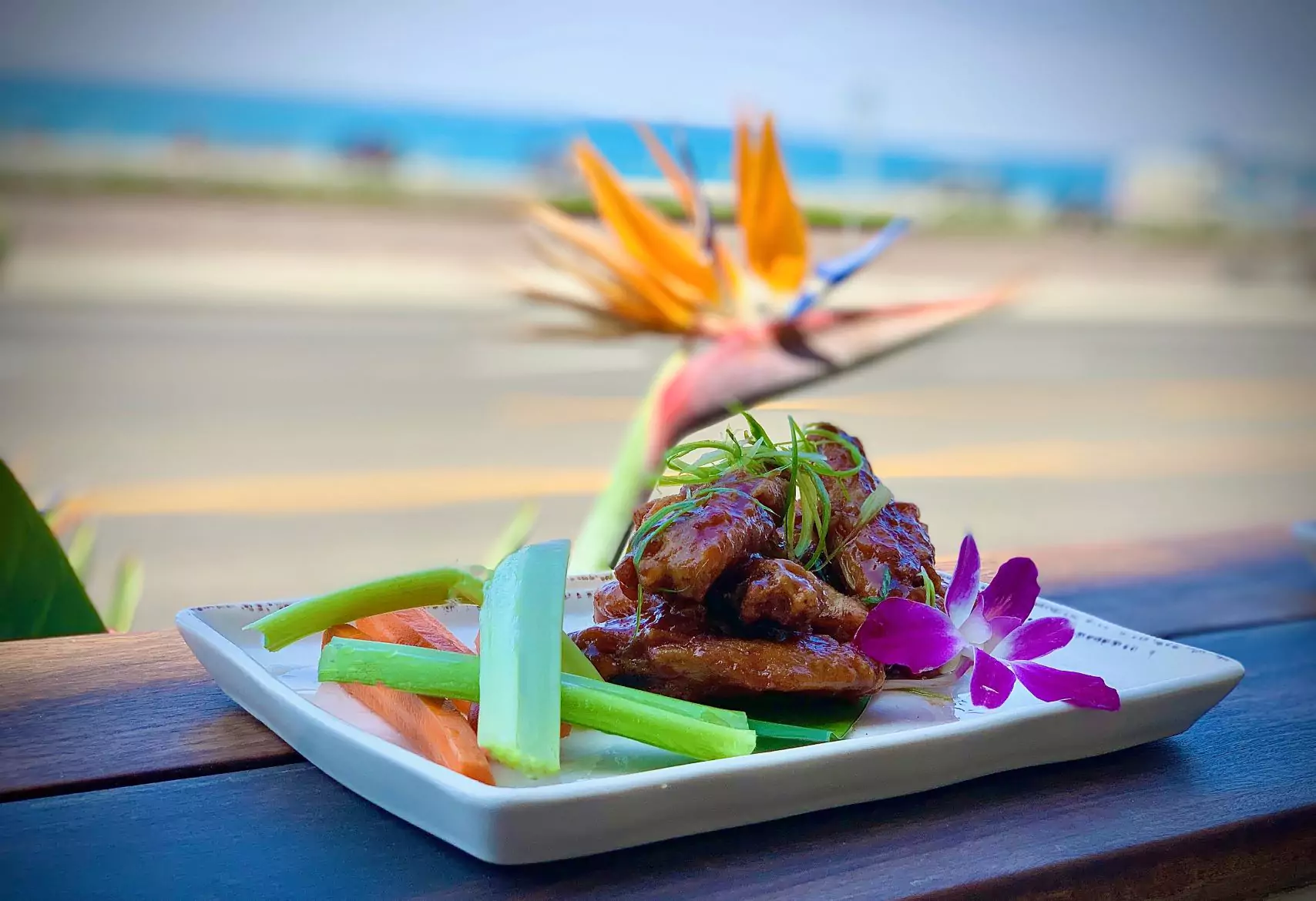 plate of wings with sauce garnishes celery and carrot sticks and edible flowers on outdoor patio dining table with views of the ocean in background