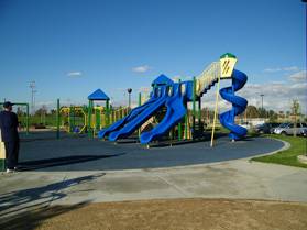 Playground with Blue, green, and yellow sets and walking sidewalks in the foreground at the Fountain Valley Recreation Center & Sports Park