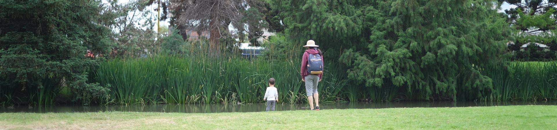 Mom wearing a maroon hoodie, straw brimmed hat, gray capri pants, and backpack with young toddler child looking at a pond and reed plants at the Fullerton Arboretum and Botanical Garden