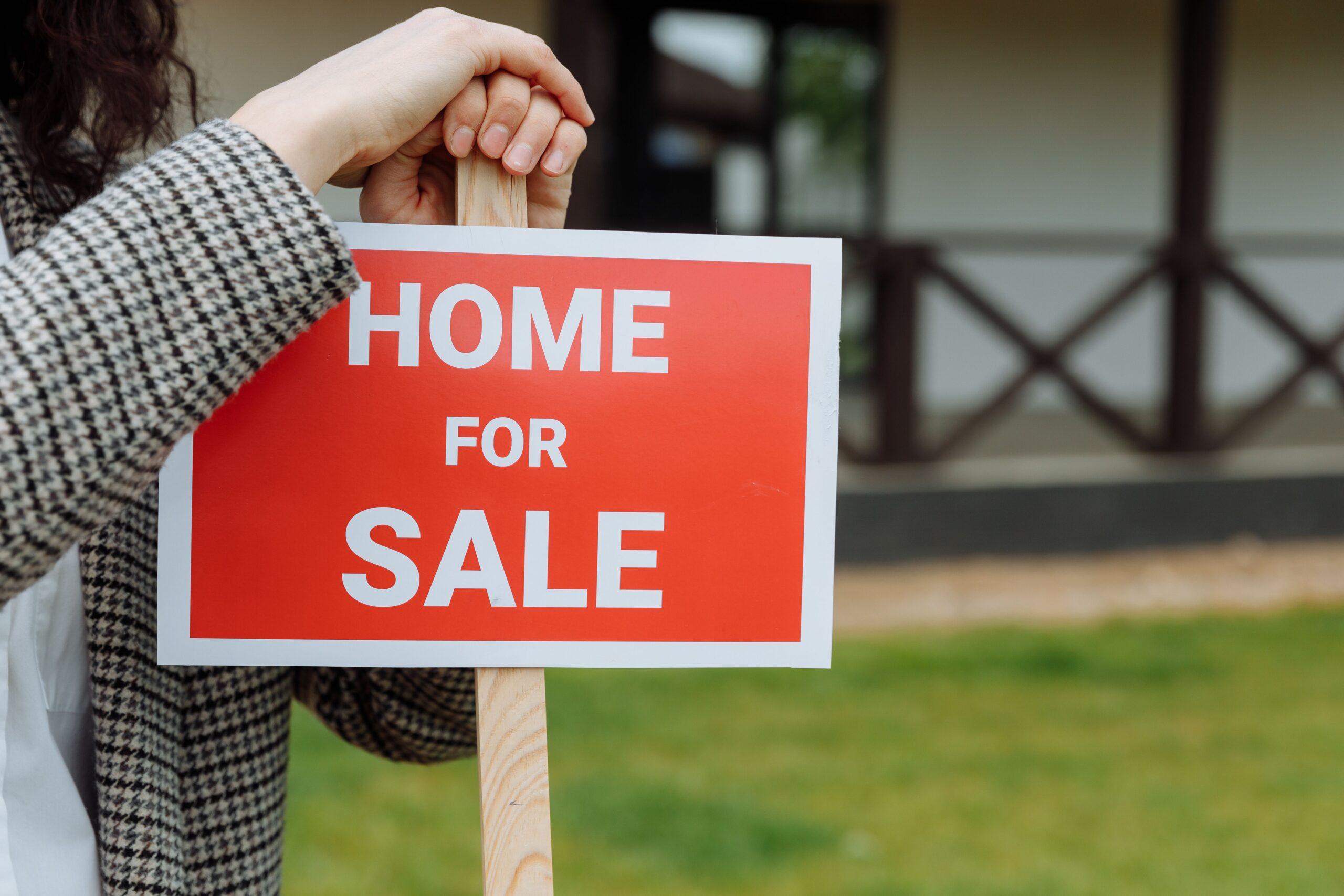 Woman holds a &ldquo;HOME FOR SALE&rdquo; sign in the ground, representing a seller&rsquo;s market in Orange County real estate
